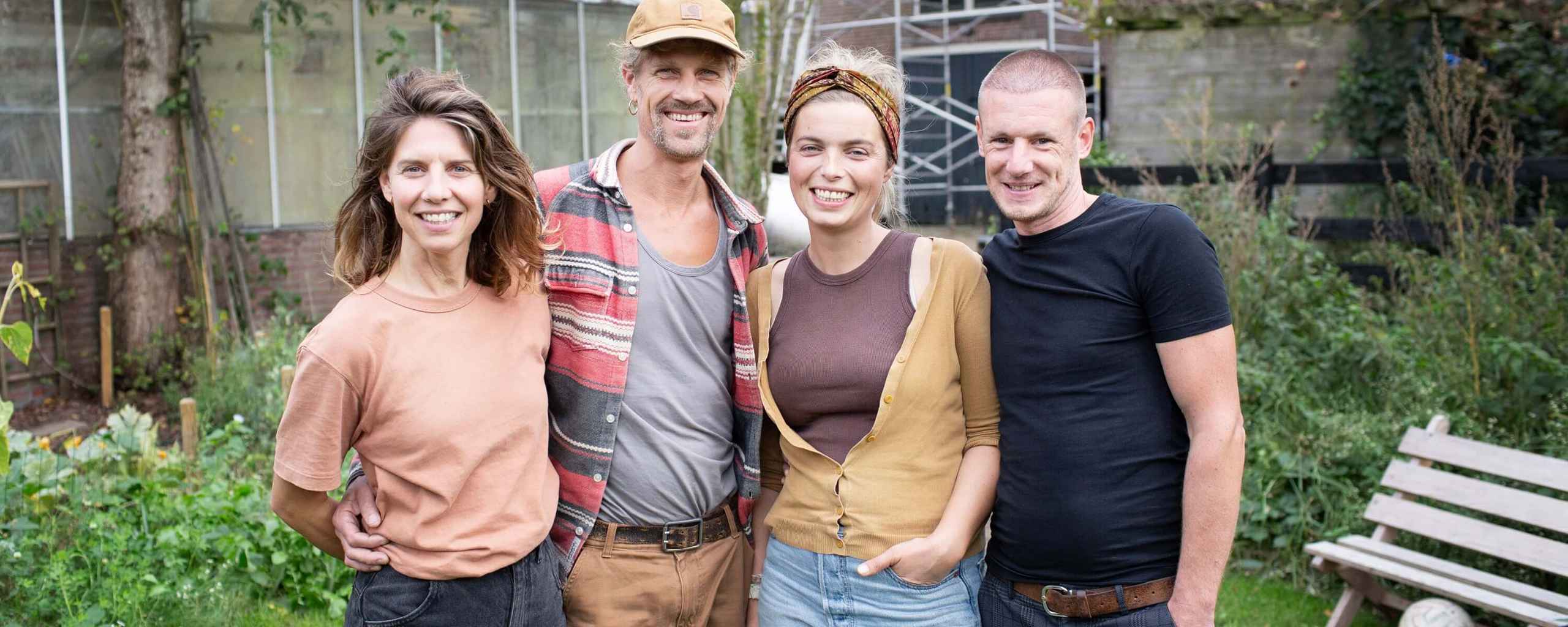 Vier mensen staan samen in een tuin. Ze hebben hun armen om elkaar heen en kijken naar de camera. Op de achtergrond staan een kas, bomen en een huis met steigers.